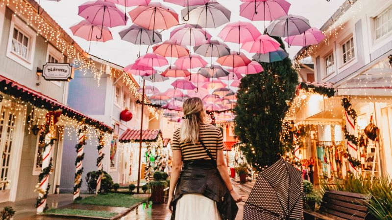 Mulher loira em penedo olhando para a rua decorada com luzes e apetrechos natalinos e com dezenas de guarda-chuvas em tons de rosa no céu.